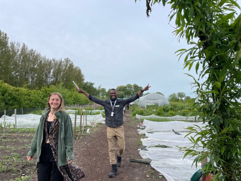 A smiling young woman and a happy young man with arms outstretched enter Alnarp's agroecology farm through a greenery covered arch