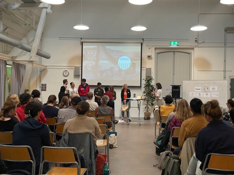 A seminar room with an audience seen from behind and 7 presenters in front